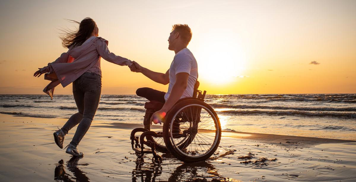 Uomo in sedia a rotelle con la fidanzata felici al tramonto sulla spiaggia - Foto di Nejron Photo da Adobe Stock