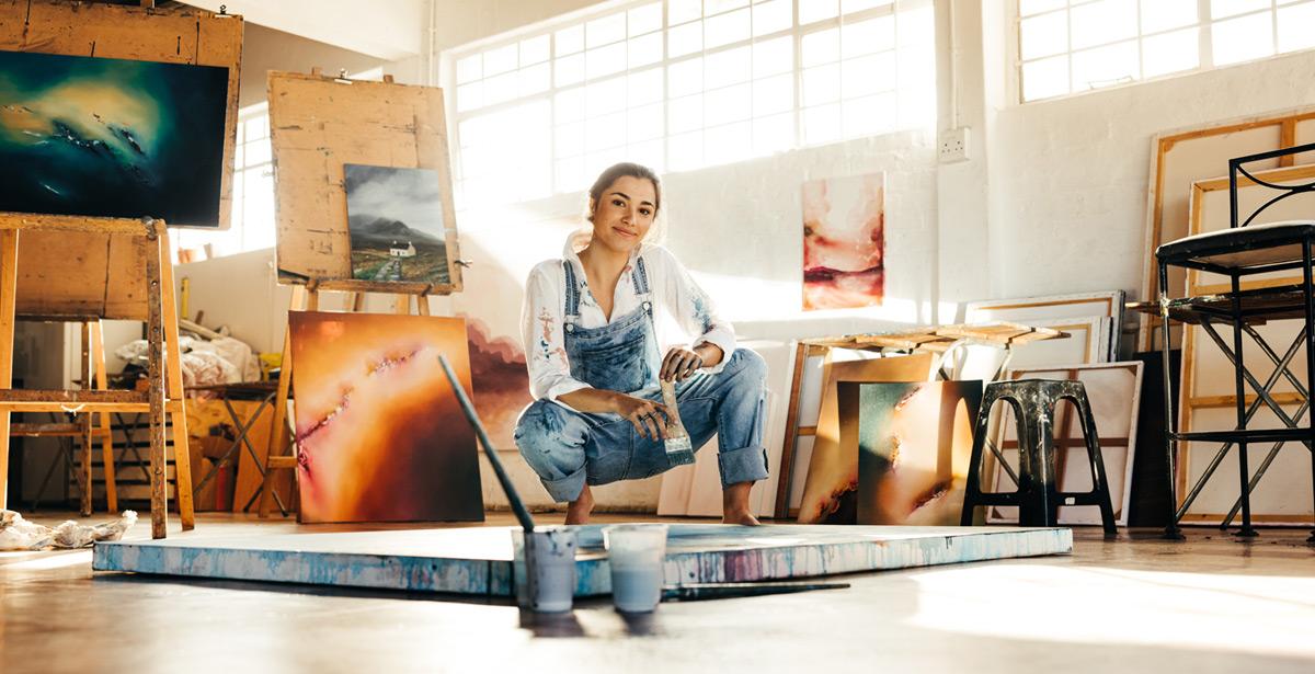 Giovane pittrice inginocchiata nel suo studio e circondata da propri dipinti - Foto di Jacob Lund da Adobe Stock