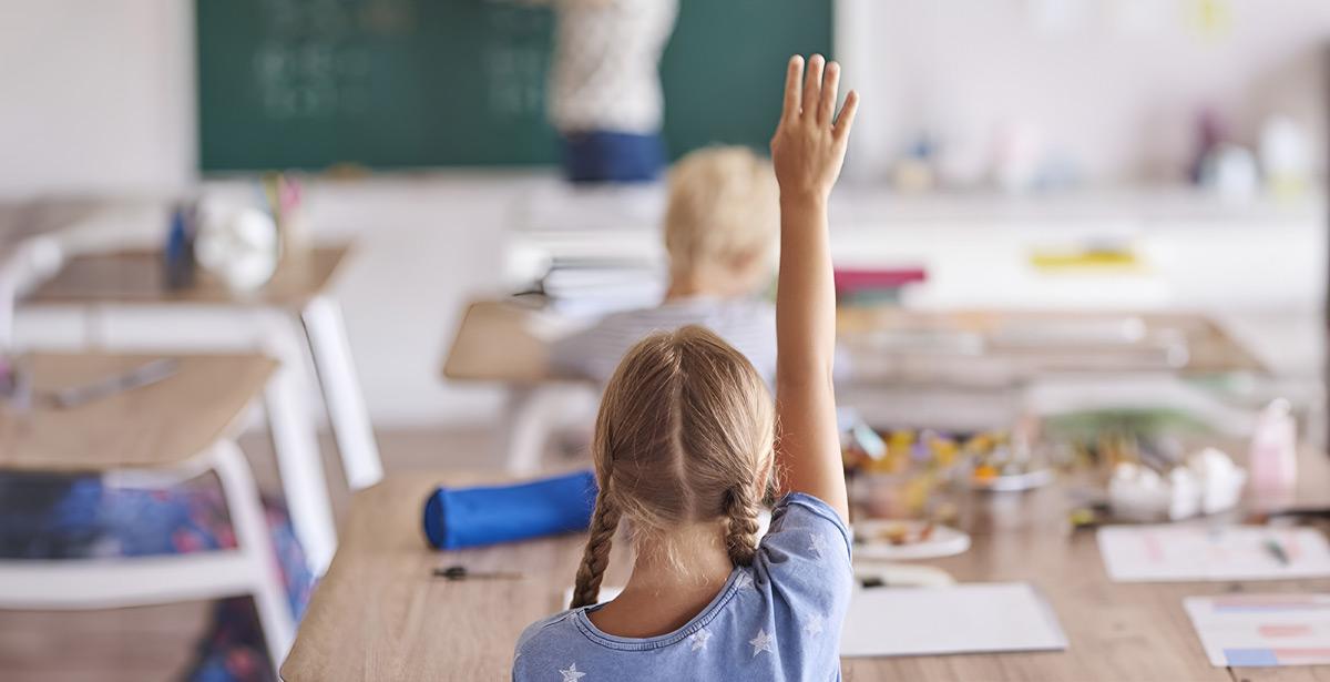 bambina con mano alzata di spalle tra i banchi di scuola